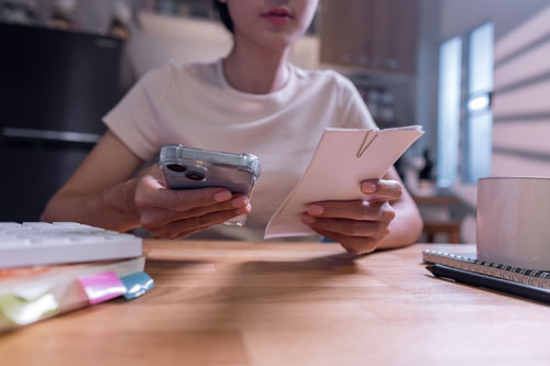 Eine Person sitzt an einem Arbeitstisch und hält ein Smartphone und Zettel in der Hand