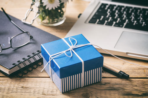 Beitragsbild zeigt ein blaues Geschenk auf einem Schreibtisch mit Laptop, Notizblock und einer Vase Gänseblümchen