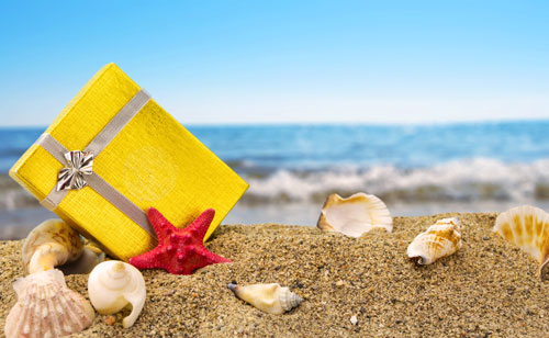 Beitragsbild zeigt ein sonnig-gelbes Geschenk auf einem Sandstrand zwischen Muscheln und dem Ozean